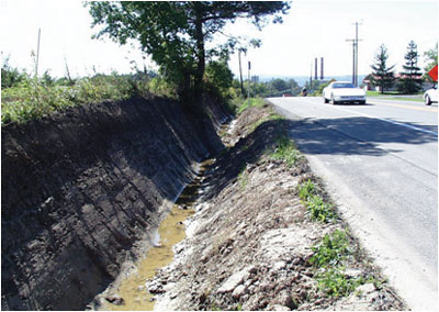 Photo. This photo shows a deep, v-shaped, muddy drainage ditch.