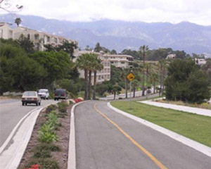 Photo.  A shared use path that runs parallel to a two lane roadway with a landscaped buffer in between.  The path is located on a hill that has a horizontal curve at the bottom.  The path has a solid yellow centerline and the climbing lane is wider than the downhill lane to allow faster cyclists, traveling uphill, to pass slower cyclists. Passing space isn’t provided for the cyclists traveling downhill, which is particularly critical given that there is a horizontal curve at the bottom of the hill.