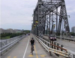 Photo.  A bridge with a pedestrian lane on the left adjacent to two bike lanes to the right.  Two travel lanes for motor vehicle traffic are to the right of the bike lanes and are separated from non-motorized traffic by the bridge supports and two guardrails.  
