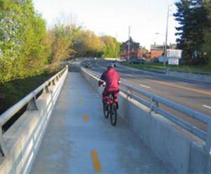 Photo.  A bridge with a shared use path on the left and the multilane roadway on the right, separated by a concrete barrier.  The path is lined with sand and road debris cast onto the cycling path from sanding and plowing operations. Sand on the pavement can cause a loss of traction for cyclists.