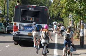 Photo.  A two lane roadway with a bus traveling in the right lane along with numerous cyclists.  A high volume of cyclists and regular bus service can create conflicts between buses and cyclists. One cyclist is riding in the bus driver’s blind spot. This condition requires vigilance from the bus operator and predictable behavior from the cyclist. 